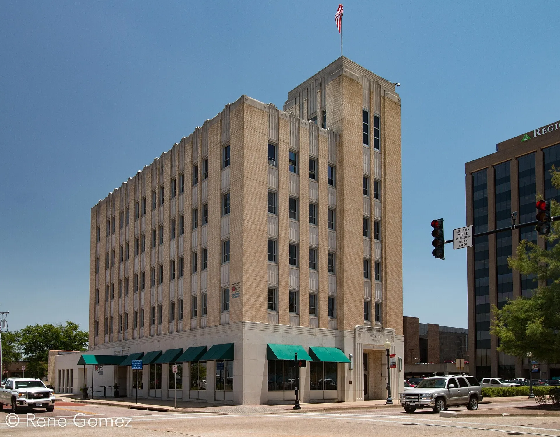 A buff brick Art Deco office building with a stepped tower and green awnings over its ground floor, at a street corner.