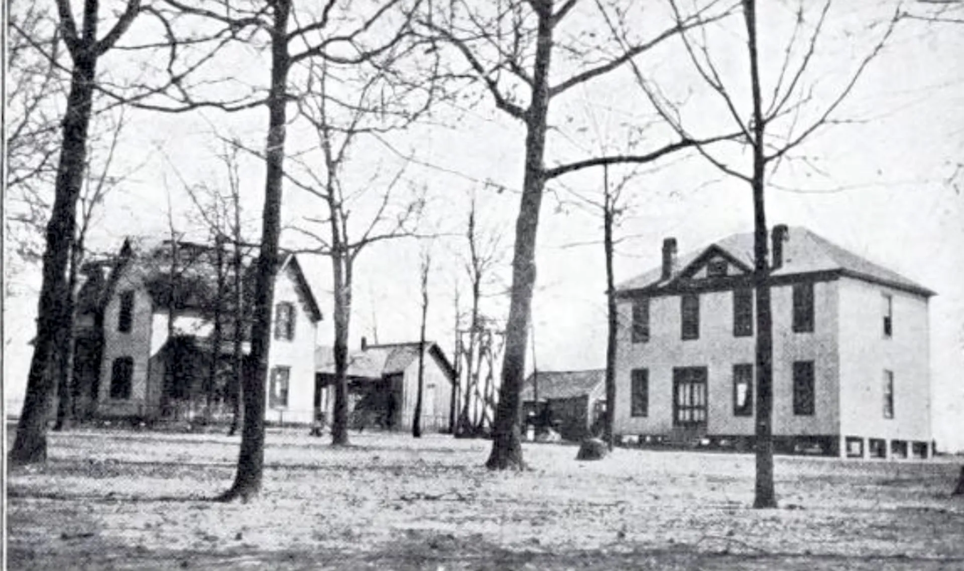 Two white frame buildings among bare trees: a two-story house and a larger two-story hall with a hipped roof.