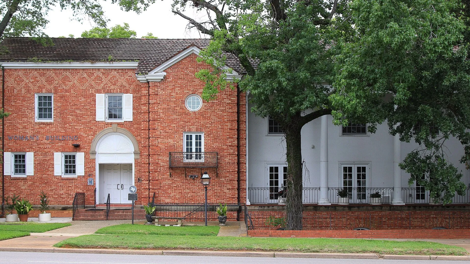 A two-story red brick building with white trim and a white-columned wing, behind a lawn and a large oak.