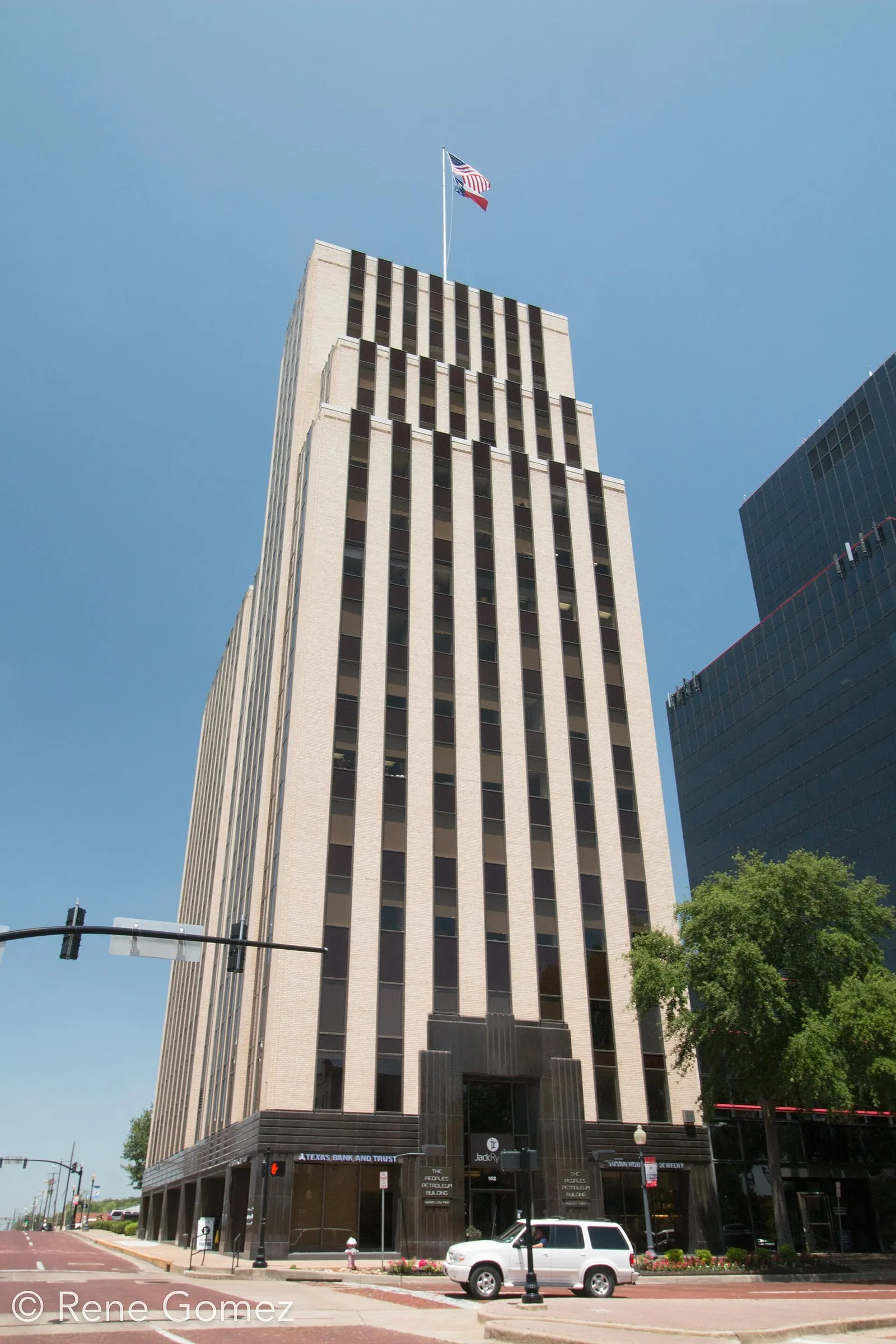 A tall pale tower striped with columns of windows and stepped back near its top, a flag on its roof, at a street corner.