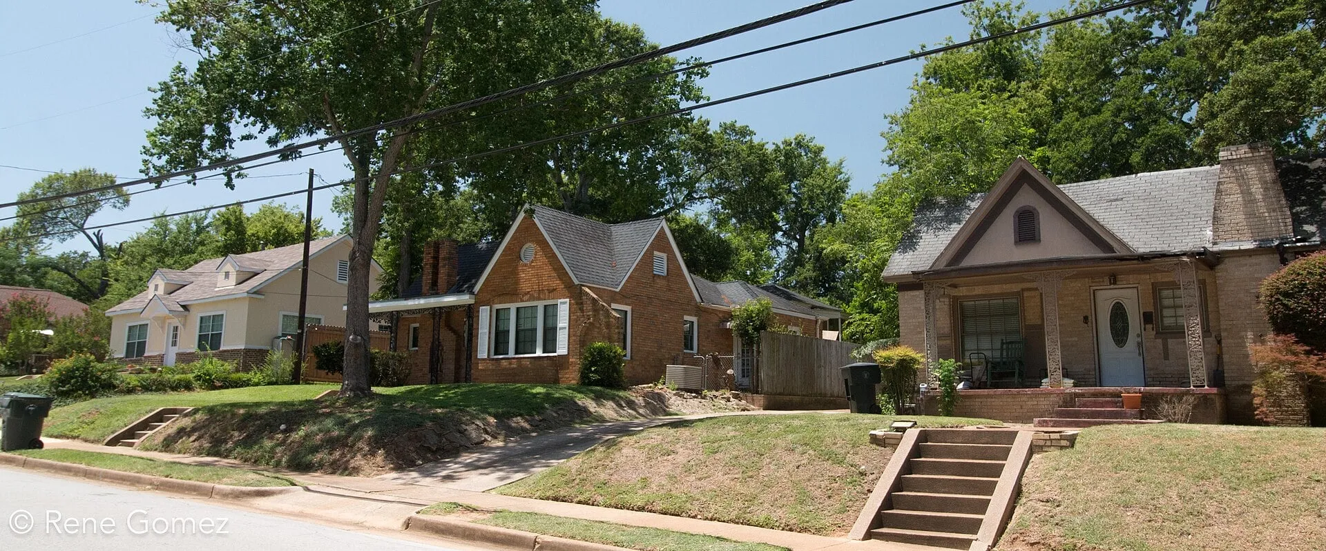 A row of brick and stone bungalows on a sloping lawn above the street, under tall trees.