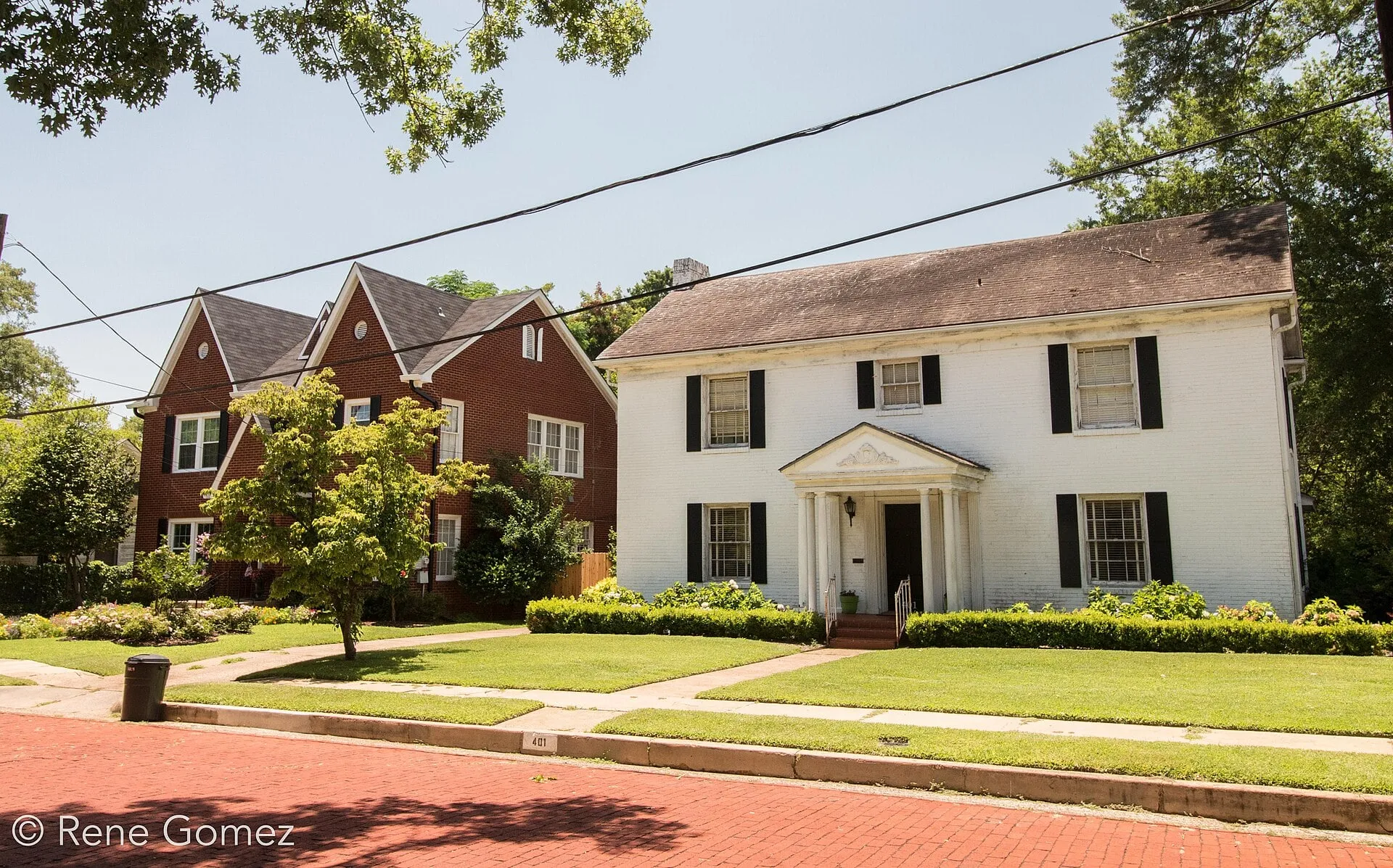 A white two-story house with black shutters and a columned porch above a red brick street, with brick houses beside it.