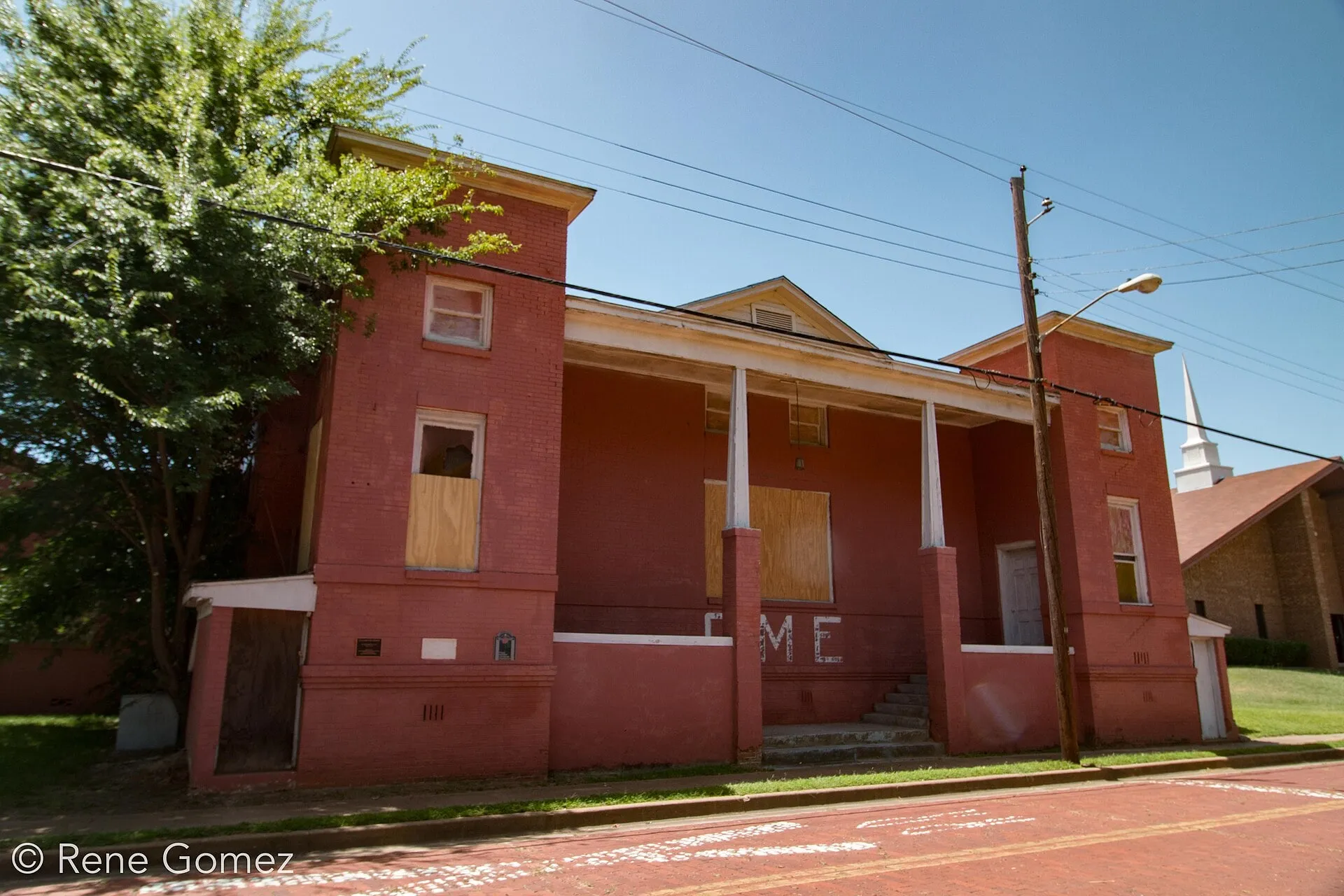A two-story church of red-painted brick with a columned porch between two square towers, its windows boarded, beside a brick street.