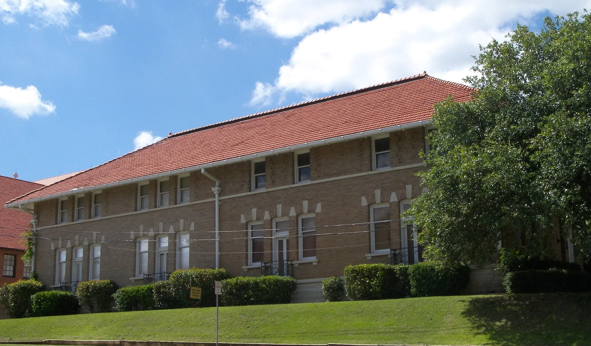 A two-story buff brick building with a red tile roof and rows of tall windows, above a sloping lawn.
