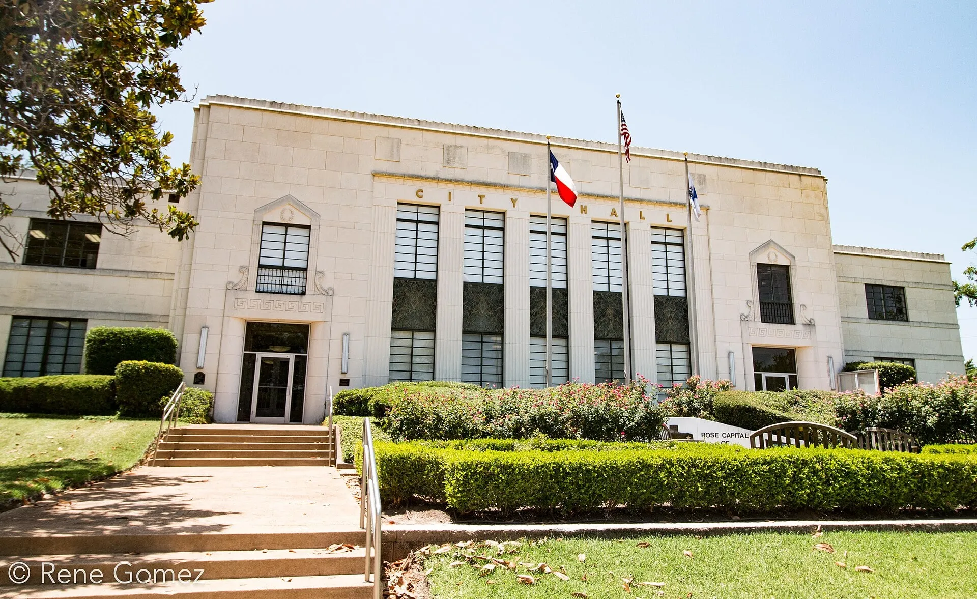 A pale stone building with tall columns of windows and the words City Hall across its front, flags beside it and steps up a hedged lawn.