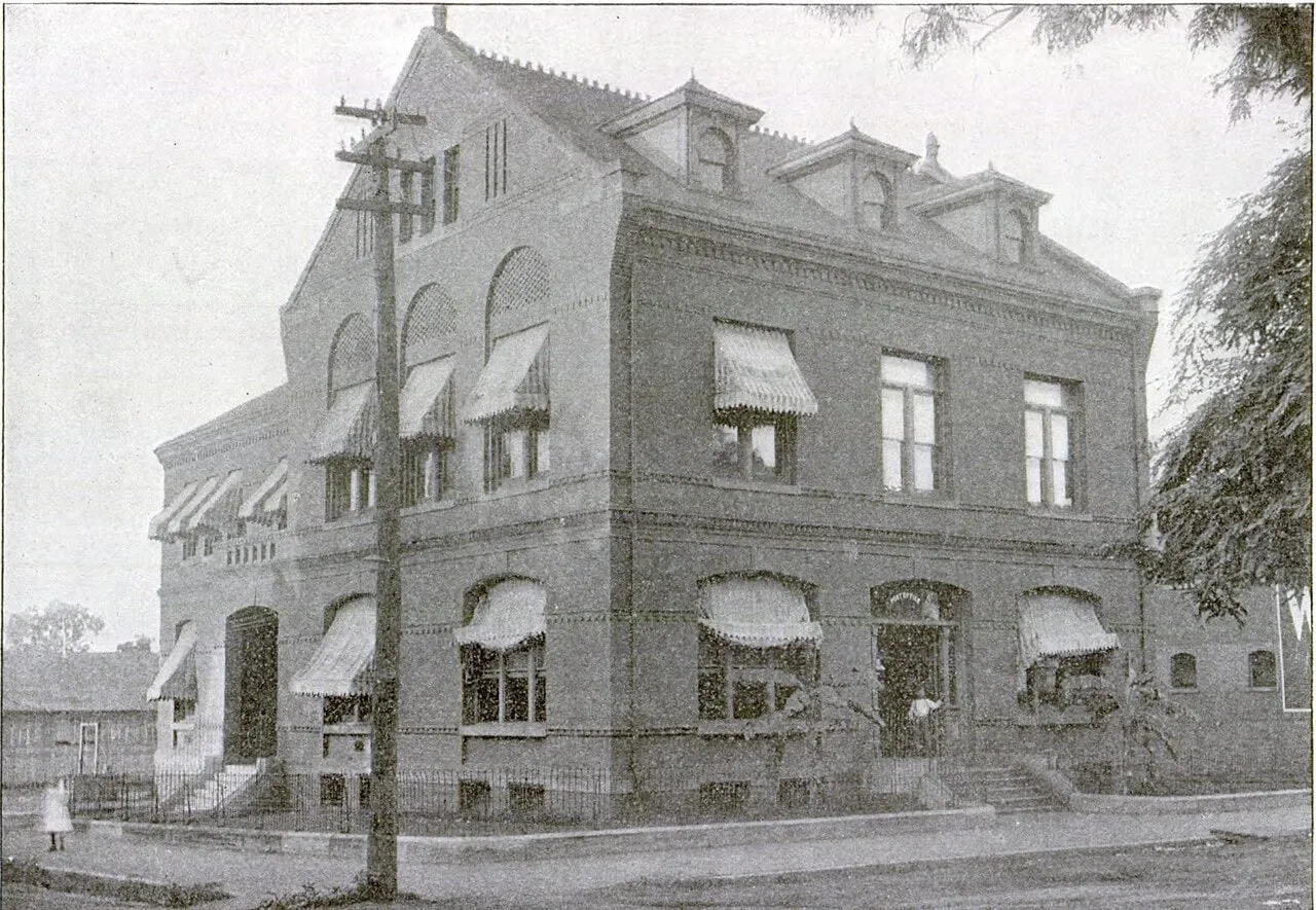 A three-story brick building with arched windows, striped awnings and dormers in its roof, on a street corner.