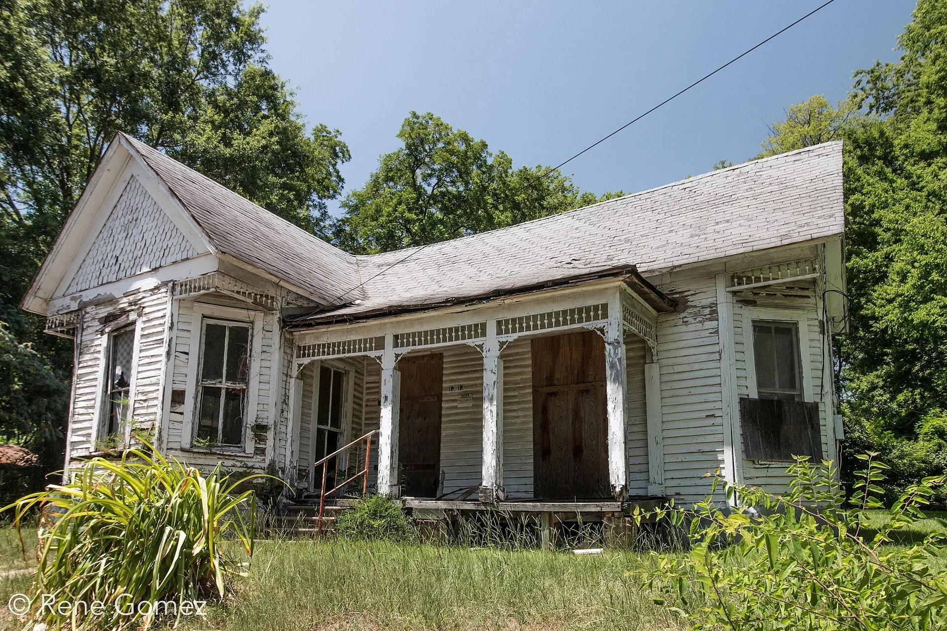A weathered one-story white frame house with a front gable and a deep porch, in tall grass under trees.